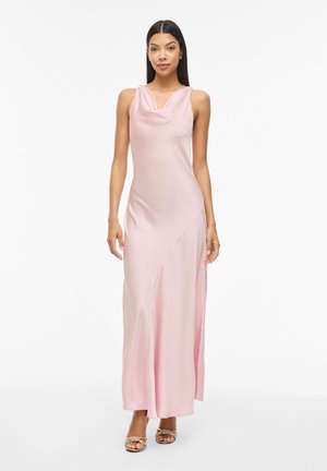 Kobieta ubrana w bezrękawową bladonóżową satynową suknię wieczorową z dekoltem typu kapturek i złote plecione sandały na odkryty palec, stojąca na białym tle.