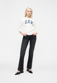 Femme blonde debout, les mains dans les poches, portant un t-shirt blanc à manches longues GAP, un jean noir à jambes droites et des chaussures à talons noires sur fond blanc.