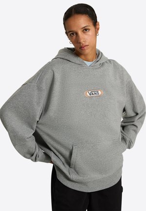 Junge Person mit dunklem Haar trägt einen lockeren grauen Vans-Hoodie und schwarze Hose, steht mit den Händen in der Hüfte vor einem einfarbigen Hintergrund.