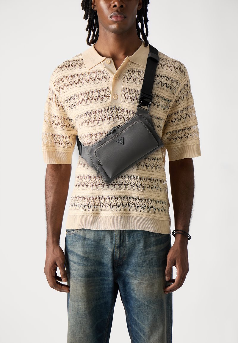 Borsa a tracolla grigia in materiale liscio con chiusura a cerniera e logo triangolare, indossata sopra un polo lavorato beige con motivo e jeans in denim.