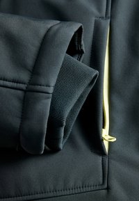Gros plan sur une veste noire mettant en avant un tissu doux, une manchette élastique verte et une fermeture éclair jaune. Détails de couture remarquables sur le matériau.