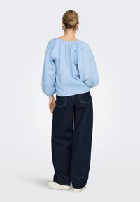 Femme aux cheveux coiffés en chignon porte des boucles d'oreilles créoles, un chemisier bleu clair à manches bouffantes, un jean large foncé et des chaussures blanches, debout, dos visible.
