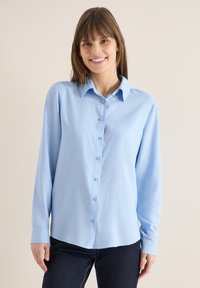Femme souriante aux cheveux bruns portant une chemise bleu clair à boutons et un pantalon foncé, debout devant un mur beige uni.