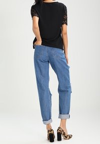 Sort top med blondeærmer, blå slidte jeans og leopardmønstrede sko med kraftige hæle. En naturlig frisure ses fra bagenden.