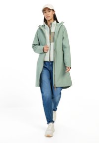 Lichtgroene waterdichte regenjas met een capuchon, met een rits aan de voorkant, gelaagd over een lichte fleece, gecombineerd met blauwe jeans en lichte sneakers.