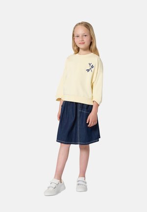 Gelber Pullover mit Libellenmotiv, kombiniert mit einem blauen Jeansrock mit seitlichen Taschen und weißen Turnschuhen. Einfaches, lässiges Outfit.