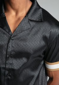Camicia nera a maniche corte con texture lucida, delicato motivo logo su tutta la superficie e accenti a strisce dorate e bianche a contrasto sulle maniche.