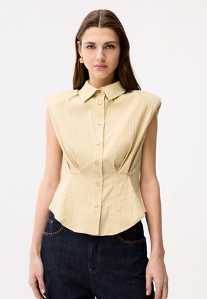 Kvinde med langt brunt hår iført gyldne øreringe i hoops, en ærmeløs beige knap-bluse med polstrede skuldre og mørkeblå jeans.