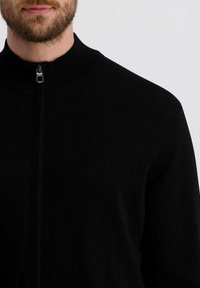 Schwarzer Reißverschlusspullover mit hohem Kragen, gestrickter Textur und silbernem Reißverschlussdetail, mit einem taillierten Design ohne sichtbare Logos oder Muster.