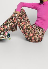 Kv ětinové rozšířené kalhoty s lesklým povrchem mají na černém pozadí živé růžové, žluté a zelené květy; doplněné dlouhým růžovým topem s dlouhým rukávem.