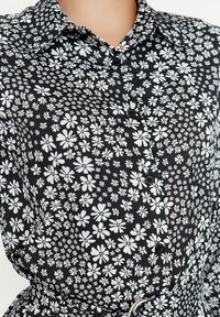 Schwarzes Hemd mit Knöpfen und einem weißen Blumenmuster. Der Stoff ist glatt mit einer leicht glänzenden Textur. Auffällige Details sind der klassische Kragen und schwarze Knöpfe.