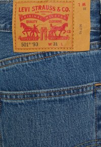 Os jeans em denim azul da Levi's apresentam um remendo retangular em pele castanha com o logótipo impresso em vermelho, número do modelo 501 '93, e detalhes de tamanho.