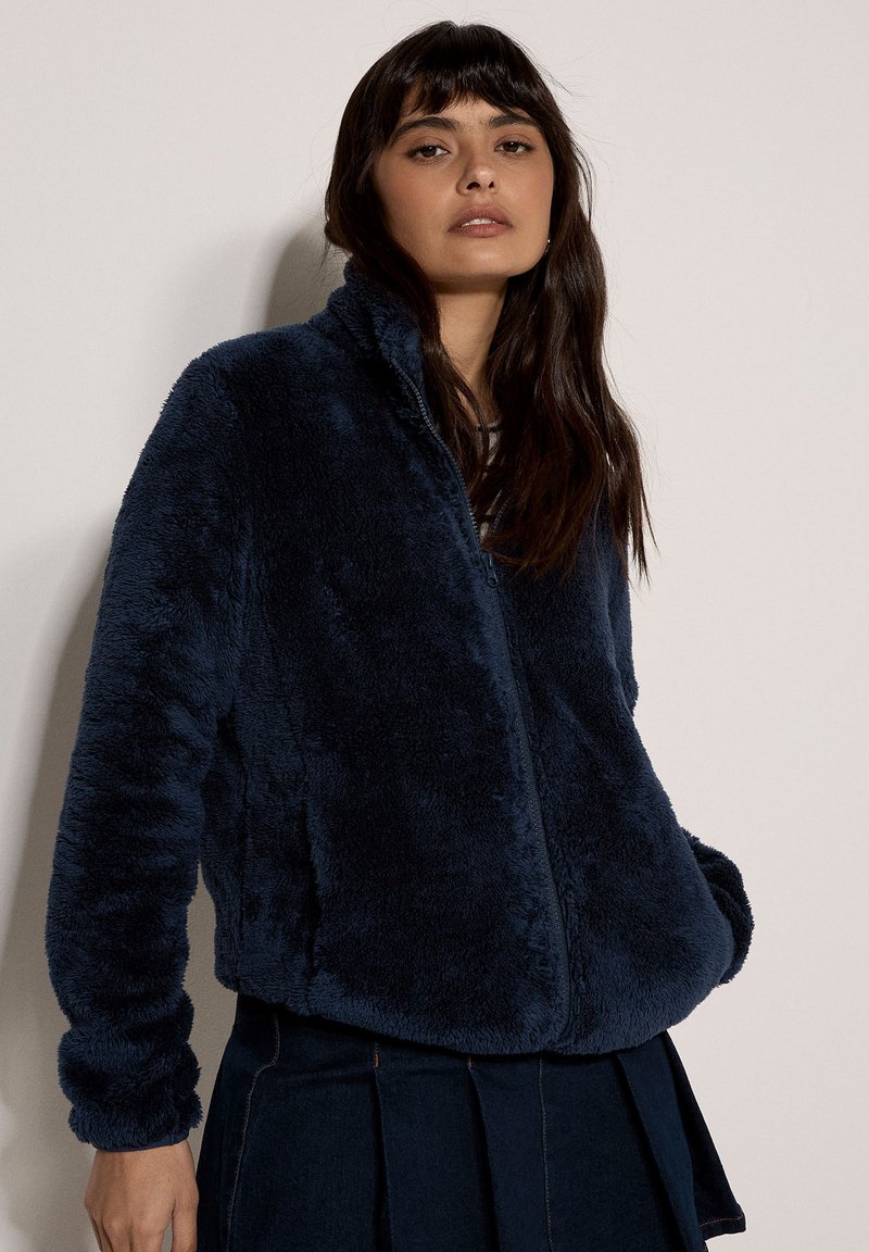 Veste en fausse fourrure bleu marine avec col montant et fermeture éclair. Comprend des manches longues et un tissu doux et texturé. Portée sur une jupe sombre.