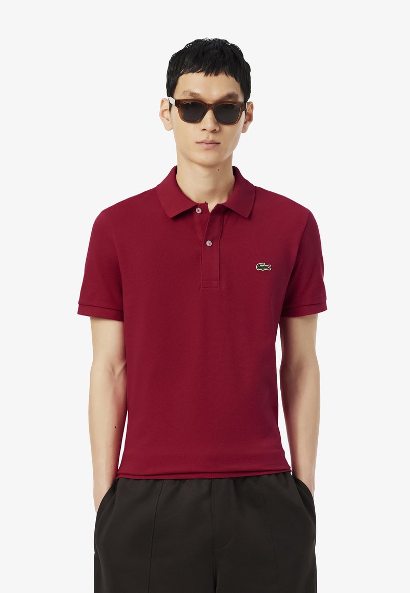 Hombre con gafas de sol marrones, camiseta polo roja con logo de cocodrilo y pantalones negros, con las manos en los bolsillos, contra un fondo blanco.
