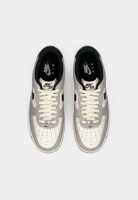 Nike Air Force 1 cipők világosszürke velúr felső résszel, fehér bőr orrrésszel, fekete pipa logóval és krémszínű fűzőkkel. Jellemzője a perforált részletek.
