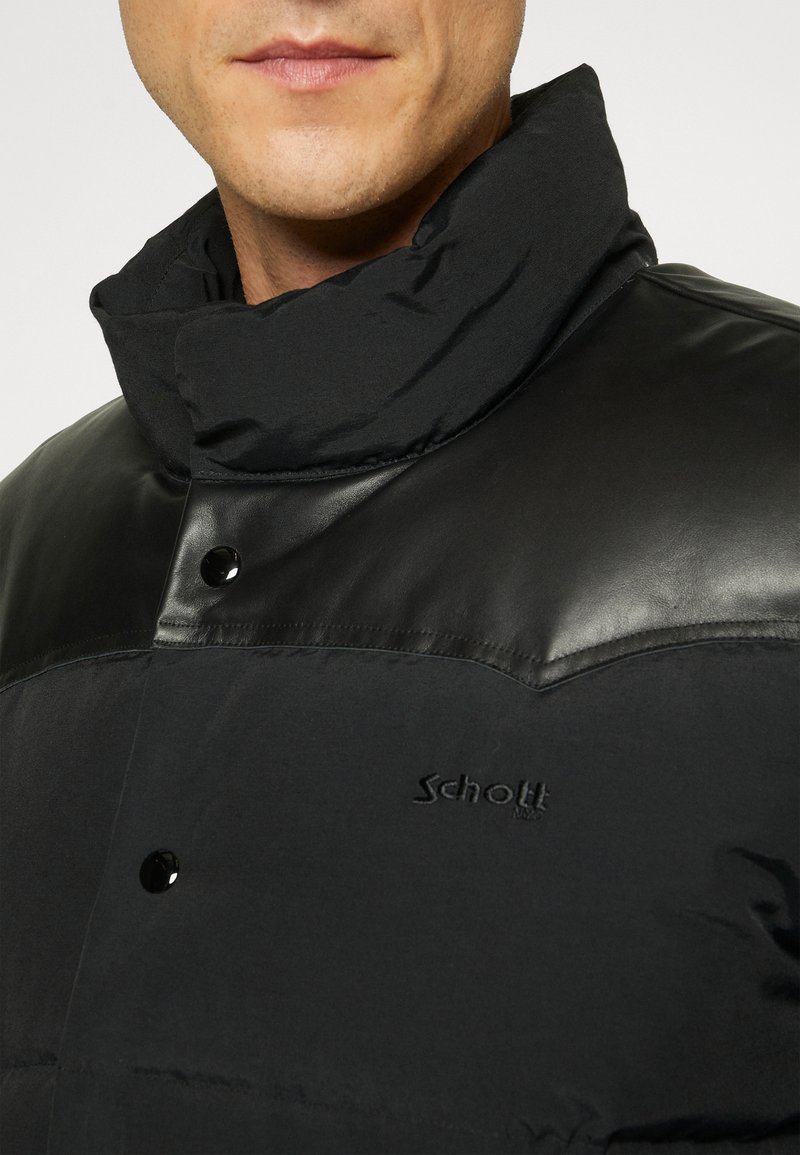Giacca nera con colletto alto, caratterizzata da una combinazione di texture opache e brillanti, chiusura a pressione e logo in rilievo sul petto.