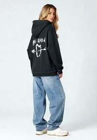 Hoodie noir oversize avec du texte graphique blanc et des flèches dans le dos, associé à un jean bleu clair ample et des baskets beiges.