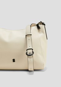 Krémová kožená kabelka přes rameno s uvolněným designem, nastavitelným popruhem a černými kovovými akcenty. Na přední straně se nachází jemné logo.