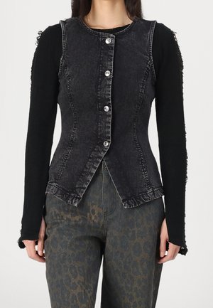 Nainen pukeutuneena istuvaan mustaan, napitettuun farkkuliiviin mustan pitkähihaisen paidan ja tummanharmaiden leopardikuvioisten housujen päällä.