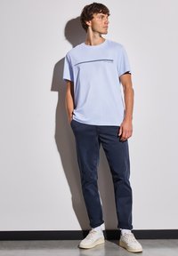 T-shirt bleu clair avec une rayure noire horizontale et un texte graphique. Associé à un chino marine et des baskets blanches, créant un look décontracté.