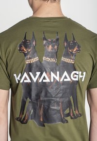 Camiseta de algodón verde oliva con un estampado gráfico de tres perros Doberman con collares con tachuelas doradas y el texto "KAVANAGH" debajo.
