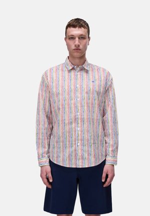 Giovane uomo che indossa una camicia a righe verticali multicolori e pantaloncini blu scuro al ginocchio, in piedi davanti a uno sfondo chiaro e uniforme.
