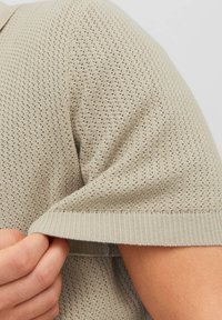 Maglione lavorato a maglia di un leggero beige con un motivo testurizzato, caratterizzato da maniche corte e orlo a coste. Il materiale sembra morbido e traspirante.