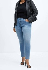 Personne portant une veste en cuir noire, un haut noir, un jean bleu clair et des sandales noires à talons hauts et à bouts ouverts, debout devant un fond uni.