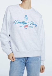 Lichtgrijze sweatshert met blauwe geborduurde tekst "Brooklyn Bridge," met een accent in de vorm van de Amerikaanse vlag. Losse pasvorm en ribgebreide manchetten.