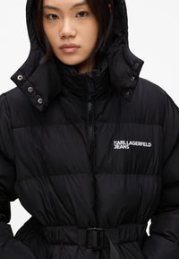 Schwarze Daunenjacke mit hohem Kragen, Reißverschluss und tailliertem Gürtel. Mit Logo-Print und Druckknopfdetails an der Kapuze.