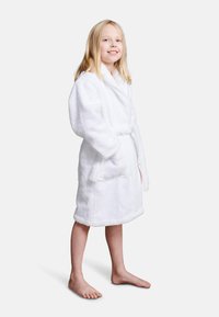 Accappatoio bianco in spugna con colletto sciallato, cintura in vita e due tasche frontali, indossato da una ragazza a piedi nudi.Texture morbida, lunghezza fino al ginocchio.