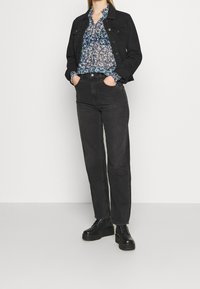 Donna che indossa una giacca in denim nera, una blusa floreale, jeans neri a vita alta e stivali neri con piattaforma e lacci, in piedi con le mani nelle tasche.