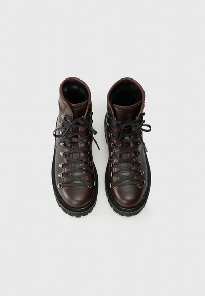 Scarponi da trekking in pelle marrone con lacci neri. Presentano occhielli argentati, una suola in gomma testurizzata e un design alto alla caviglia per un maggiore supporto.