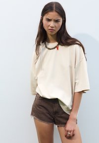 Cremefarbenes Oversized-T-Shirt aus weicher Baumwolle, kombiniert mit braunen Jeansshorts mit ausgefranstem Saum. Accessoires mit einer dicken Kettenhalskette.