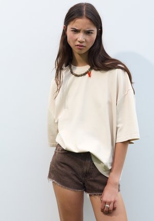 T-shirt crème surdimensionné en coton doux, associé à un short en denim marron avec un ourlet effiloché. Accessoirisé avec un collier en chaîne épais.