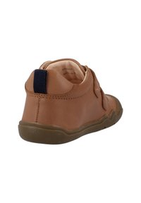 Zapato de cuero marrón con una superficie texturizada, punta redondeada y una correa de velcro. Presenta un lazo azul en la parte trasera y una suela de goma flexible.