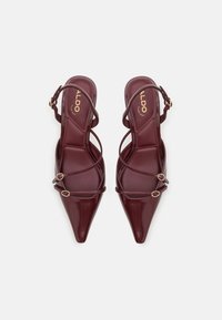 Talons slingback en cuir verni bourgogne avec un bout pointu, des brides croisées avec des accents métalliques ronds, et une semelle intérieure rembourrée.