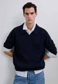 Suéter de punto navy con un cuello amplio, llevado sobre una camisa de rayas azul claro. Las mangas están arremangadas y el corte es holgado.
