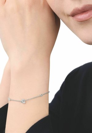 Wrist with silver chain bracelet featuring a small heart charm, resting near a face wearing a black long-sleeve top.