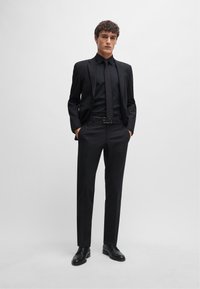 Completo nero su misura, camicia abbottonata abbinata e cravatta. Pantaloni slim-fit e scarpe di pelle nere. Texture liscia e design minimalista.