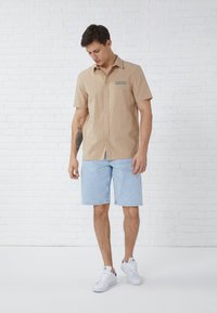 Beige short-sleeve shirt with a collar and button closure, paired with light blue denim shorts. The outfit is completed with white sneakers.