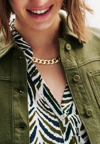 Veste en denim verte sur une chemise blanche avec des motifs abstraits verts et navy ; collier en chaîne dorée visible au niveau du cou.