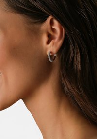 Silberne Creolen mit einem geschwungenen Design, die entlang der Vorderkante kleine klare Steine und eine polierte Oberfläche aufweisen.