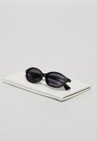 Black sunglasses with oval lenses, plastic frame, and gold accents, displayed on a white case. The lenses are dark with a smooth finish.