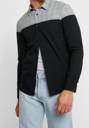 Chemise noire à manches longues avec un panneau à carreaux gris en haut, fermeture à boutons, et texture lisse. Portée avec un jean bleu clair.