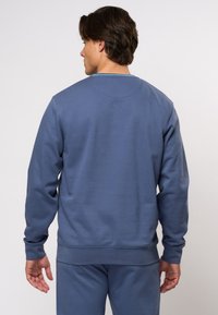 Modra trenerka s okroglim izrezom, dolgimi rokavi in ribastimi manšetami. Tkanina deluje mehko, z gladko teksturo in minimalnimi šivi.