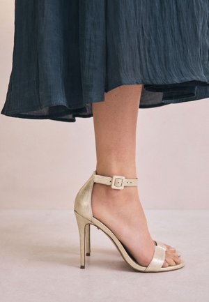 Guld højhælet sandal med tekstureret finish, åben tå og ankelrem med spænde. Bruges sammen med en flydende mørkeblå kjole.