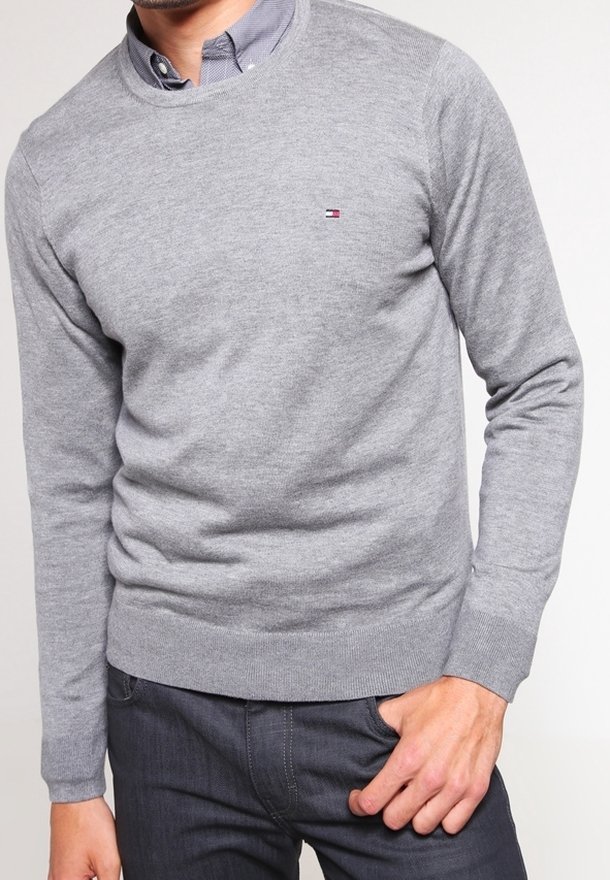 Homme portant un pull gris clair à col rond par-dessus une chemise bleue boutonnée, un pantalon gris foncé et une main dans la poche.