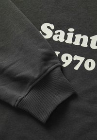 Šedá mikina s vroubkovaným manžetem, na které je výrazný bílý text "Saint 1970". Látka vypadá měkká a má hladkou texturu.