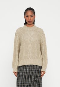 Beige kábeltört mintás pulóver magas nyakkivágással, esett vállakkal és texturált mintákkal. Fekete-fehér mintás szoknya felett viselve.